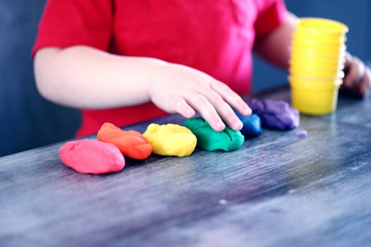 A young child with rainbow-colored modeling clay and stacking cups on a table indoors.